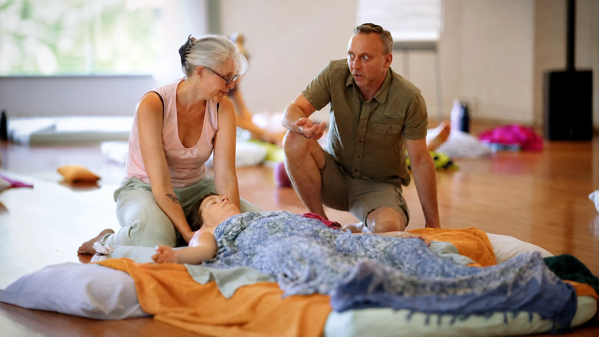 Deux adultes assis auprès d'une personne allongée sous une couverture, lumière douce venant d'une grande fenêtre
